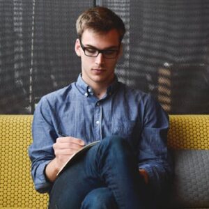 A young man in glasses writes in a notebook while sitting on a stylish couch indoors.